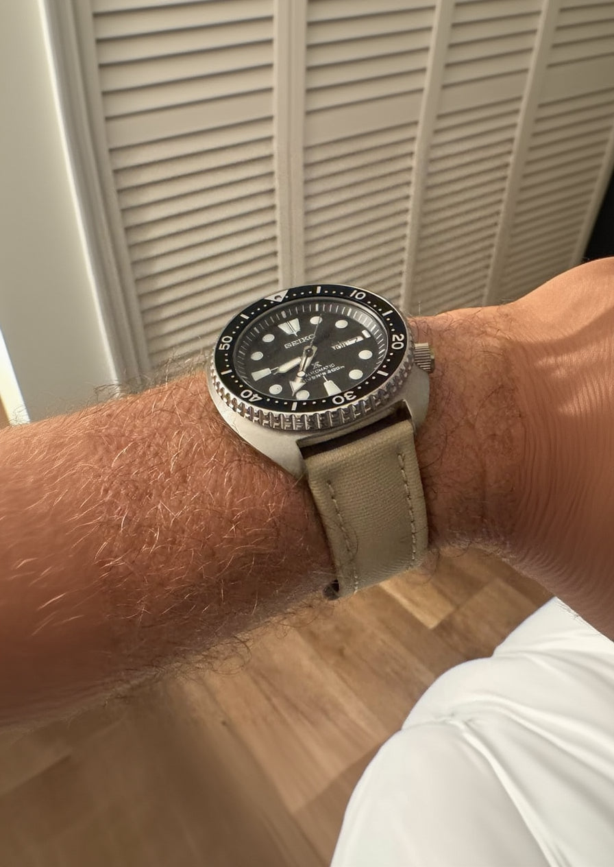 Wrist wearing a watch with a beige strap against a neutral background