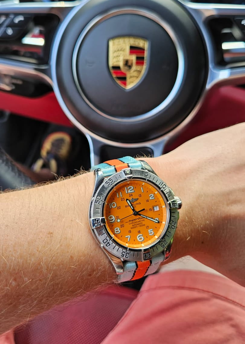Person wearing a watch with an orange face and colorful strap in front of a car's steering wheel emblem.